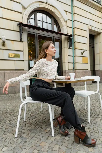 Mujer de moda con top de encaje y pantalones mirando hacia otro lado mientras sostiene el café y se sienta al aire libre - foto de stock