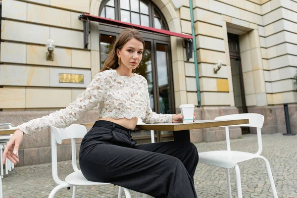 Junge Frau in Spitzenoberteil und Hose hält Kaffee und sitzt am Tisch eines Cafés in Berlin — Stockfoto