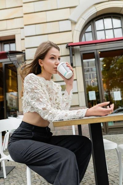 Giovane donna che beve caffè per andare mentre trascorre del tempo a tavola del caffè all'aperto — Foto stock
