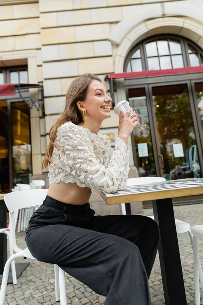 Joven alegre con top de encaje y pantalones de cintura alta sosteniendo café en Berlín, Alemania - foto de stock