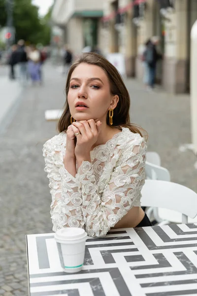 Portrait of fashionable and pretty young woman in white lace top sitting near coffee to go on table — Stock Photo