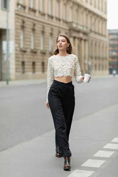 Elegante donna in pizzo superiore e pantaloni a vita alta che tengono il caffè per andare in strada a Berlino, Germania — Foto stock