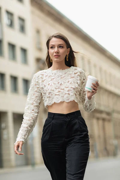 Mulher confiante em top de renda e calças de cintura alta segurando café para ir na rua em Berlim, Alemanha — Fotografia de Stock