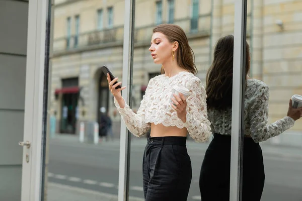 Trendige junge Frau nutzt Smartphone und hält Kaffee in der Nähe eines Gebäudes in Berlin — Stockfoto