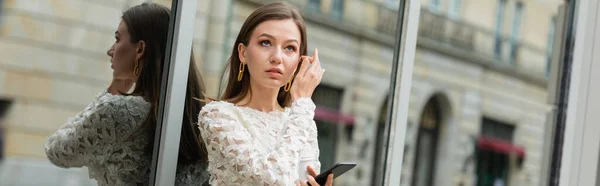 Задумчивая молодая женщина держит смартфон, стоя возле здания на улице в Берлине, баннер — стоковое фото