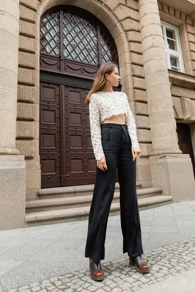Trendy woman in lace top, high waist pants and boots looking away on urban street in Berlin — Stock Photo