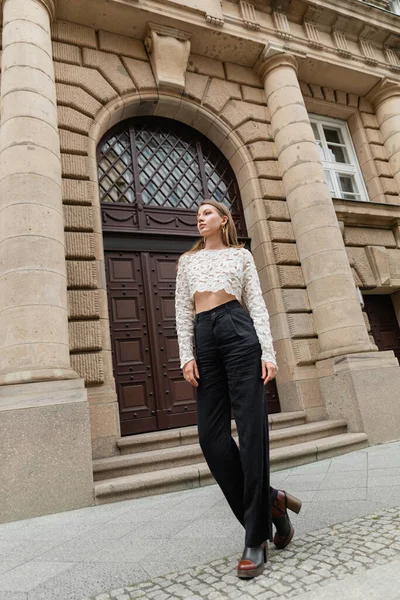 Mulher elegante confiante em top de renda e calças de cintura alta olhando para longe na rua urbana em Berlim — Fotografia de Stock