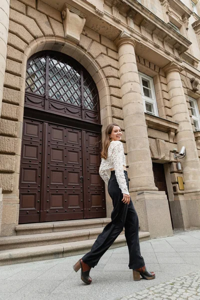 Радісна молода жінка в мереживній верхній і високій талії штани дивиться на камеру на міській вулиці в Берліні — стокове фото