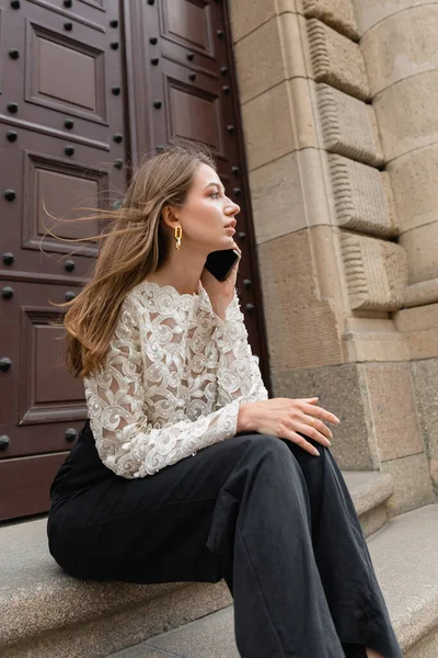 Жінка в мереживі зверху і штани розмовляє на смартфоні і дивиться далеко, сидячи на сходах біля дверей — стокове фото