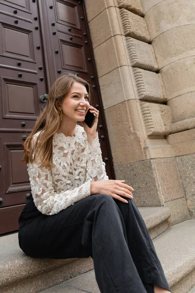 Mujer alegre hablando en el teléfono inteligente mientras está sentado en las escaleras cerca de la entrada del edificio en Berlín - foto de stock