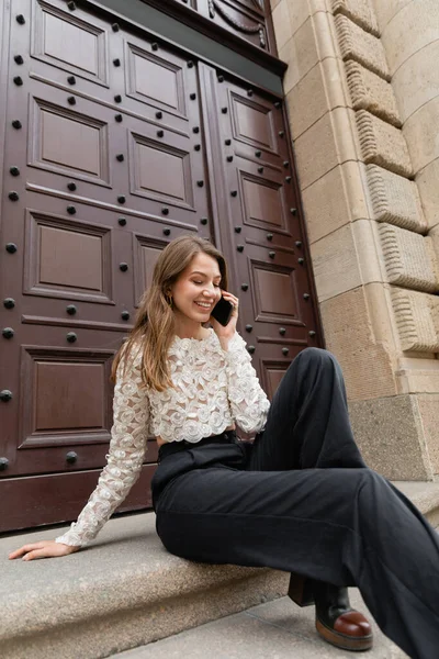 Fröhliche Frau, die auf einer Treppe in der Nähe des Hauseingangs in Berlin mit dem Smartphone spricht — Stockfoto