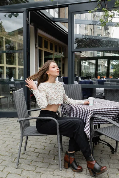 Стильная женщина трогает волосы и держит кофе, чтобы пойти, сидя на террасе открытого кафе — стоковое фото