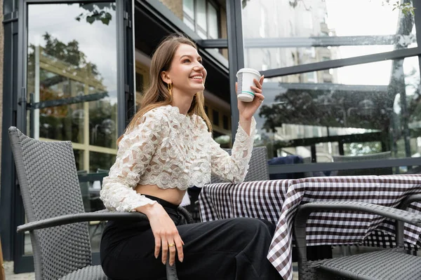 Mujer joven positiva sosteniendo café para ir mientras está sentado en la mesa en la terraza cerca de la cafetería en Berlín - foto de stock