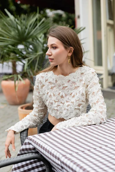 Модная молодая женщина в белом кружевном топе смотрит в сторону, сидя за столом на террасе — стоковое фото