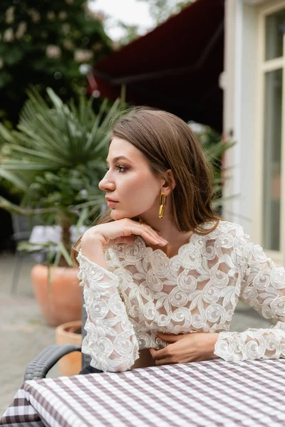Stylish woman in white lace top looking away while sitting near table on terrace of cafe — Stock Photo