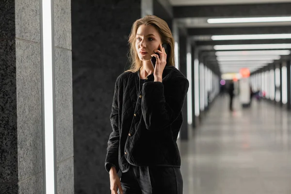 Mujer con estilo en chaqueta negra hablando en el teléfono inteligente, mientras que de pie dentro del edificio moderno - foto de stock