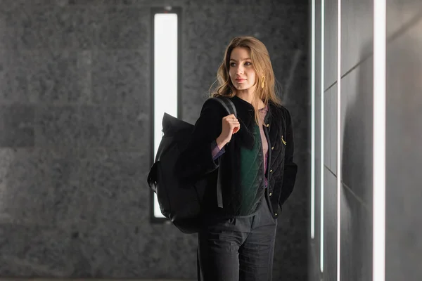 Mulher na moda em jaqueta preta segurando mochila e olhando para longe no edifício moderno — Fotografia de Stock
