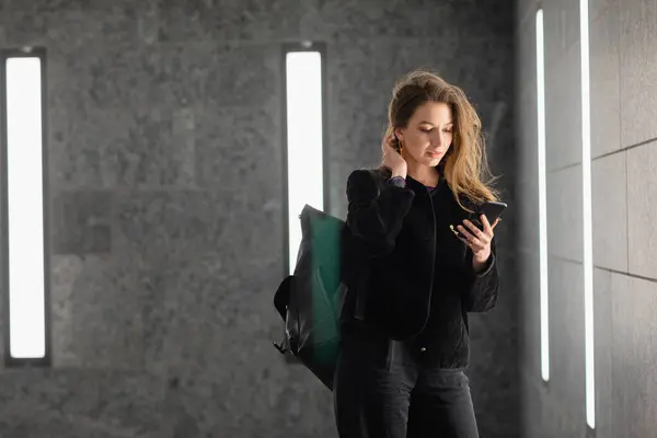 Jovem elegante com mochila usando smartphone enquanto está dentro do edifício moderno — Fotografia de Stock