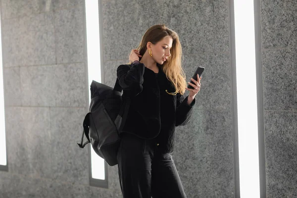 Femme élégante en veste avec sac à dos toucher les cheveux et en utilisant un smartphone près des lampes fluorescentes — Photo de stock