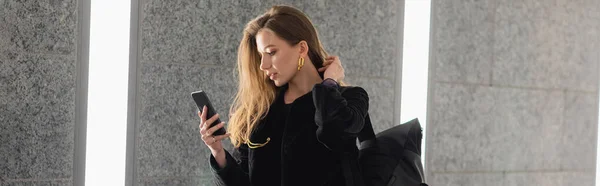 Модна жінка в куртці з рюкзаком, використовуючи мобільний телефон, стоячи біля сірої бетонної стіни, банер — стокове фото