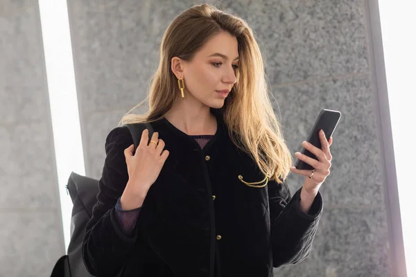 Junge Frau in schwarzer Jacke mit Rucksack und Smartphone in der Nähe von Leuchtstofflampen — Stockfoto