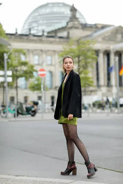 Piena lunghezza di donna alla moda in abito di seta verde, stivali e giacca guardando lontano all'aperto — Foto stock