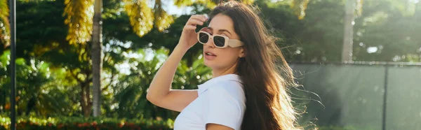 Mujer morena cautivadora y joven con el pelo largo de pie en camiseta blanca y gafas de sol de moda cerca de palmeras borrosas y verdes en Miami, día soleado, viajes, vacaciones, pancarta — Stock Photo
