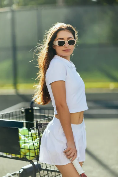 Pista de tenis en Miami, mirada enfocada, mujer joven atlética con el pelo largo de pie en traje blanco y gafas de sol mientras sostiene raqueta cerca del carrito de la pelota de tenis y fondo borroso, ciudad icónica - foto de stock