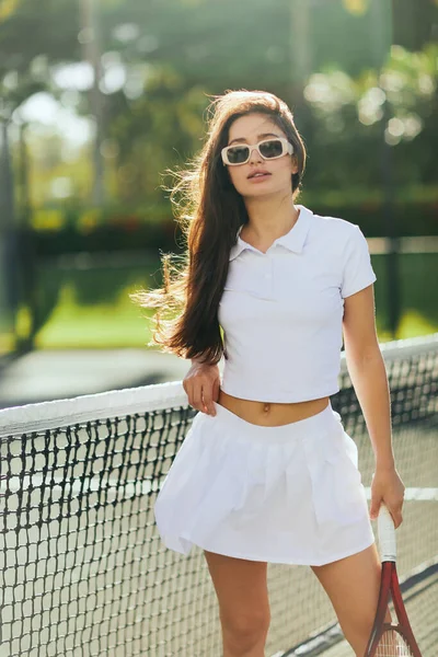 Tennisplatz in Miami, brünette junge Frau im weißen Outfit mit Tennisrock und Sonnenbrille, Schläger in der Nähe des Tennisnetzes, verschwommener Hintergrund, ikonische Stadt, Blick in die Kamera — Stockfoto
