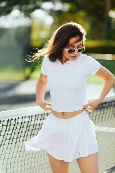 Portrait of pretty young woman with brunette long hair standing in white outfit and sunglasses near tennis net, blurred background, wind, tennis court in Miami, iconic city, female player, Florida — Stock Photo