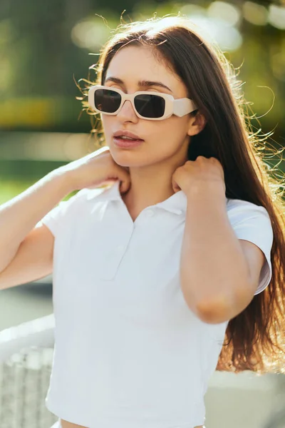 Porträt einer stilvollen jungen Frau mit brünetten langen Haaren, die in weißem Outfit und Sonnenbrille neben verschwommenem Tennisnetz auf dem Hintergrund steht, Tennisplatz in Miami, ikonische Stadt, Florida, sonniger Tag — Stockfoto