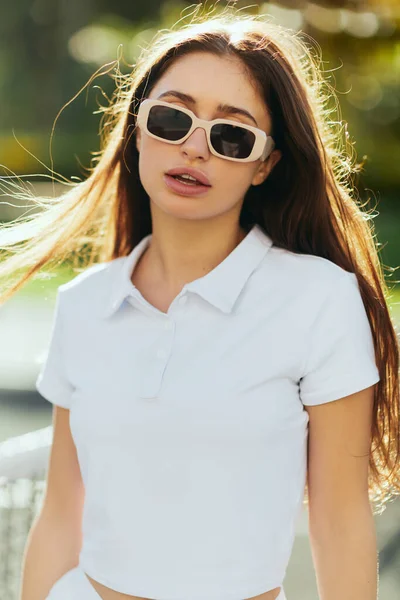Portrait of charming young woman with brunette long hair standing in white polo shirt and sunglasses near tennis net, blurred background, wind, tennis court in Miami, iconic city, Florida, sunny day — Stock Photo