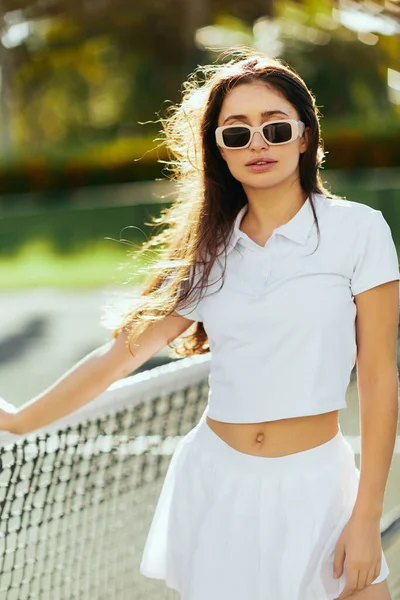 Portrait de jolie jeune femme aux cheveux longs bruns debout en tenue blanche et lunettes de soleil près du filet de tennis, fond flou, court de tennis à Miami, Floride, ville emblématique, journée ensoleillée — Photo de stock