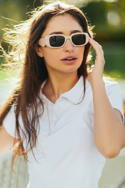 Retrato de jovem elegante com cabelos longos morena em pé em roupa branca e óculos de sol perto de rede de tênis borrada no fundo, quadra de tênis em Miami, Flórida, cidade icônica, dia ensolarado — Fotografia de Stock