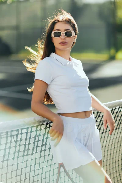 Miami, Florida, estilo de vida activo, hermosa mujer joven de pie en traje elegante y gafas de sol mientras sostiene raqueta cerca de la red de tenis, fondo borroso, ciudad icónica, día soleado, vacaciones - foto de stock