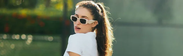 Flerte e sedutora jovem com cabelo morena em rabo de cavalo e óculos de sol em pé em camisa de pólo branco e salientando a língua no campo de ténis com fundo borrado, banner, Miami, Florida — Fotografia de Stock