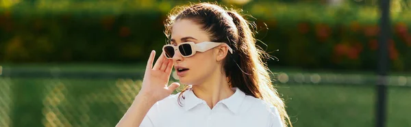 Hübsche Frau mit brünetten Haaren im Pferdeschwanz trägt weißes Outfit mit Poloshirt und Sonnenbrille, während sie auf dem Tennisplatz mit verschwommenem Hintergrund wegschaut, Miami, Florida, Banner — Stockfoto