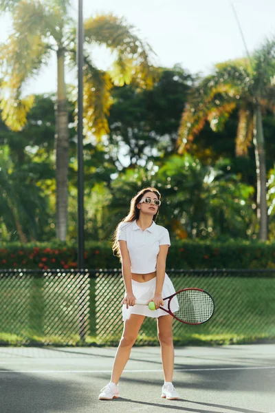 Mulher atlética morena com cabelos longos em pé em roupas brancas esportivas e segurando raquete com bola no campo de tênis em Miami, Flórida, Dia ensolarado, palmeiras no fundo borrado — Fotografia de Stock