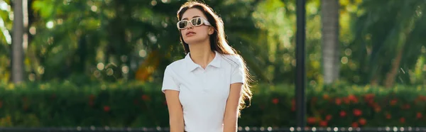 Attractive and young brunette woman with long hair standing in white polo shirt and trendy sunglasses near blurred and green palm trees in Miami, iconic city, sunny day, travel, vacation, banner — Stock Photo