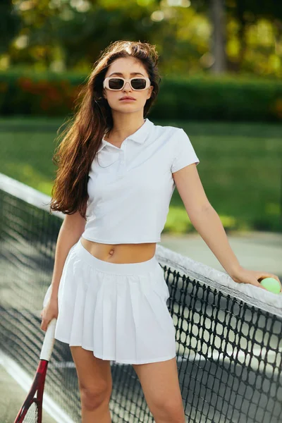 Tennisplatz in Miami, athletische junge Frau mit langen Haaren im weißen Outfit und Sonnenbrille, Schläger und Ball in der Nähe des Tennisnetzes, verschwommener Hintergrund, ikonische Stadt, Blick in die Kamera — Stockfoto