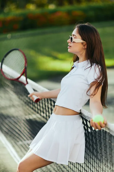 Court de tennis à Miami, jeune femme sportive aux cheveux longs debout en tenue blanche et lunettes de soleil tout en tenant raquette floue et balle et appuyé sur un filet de tennis, fond vert, ville emblématique — Photo de stock