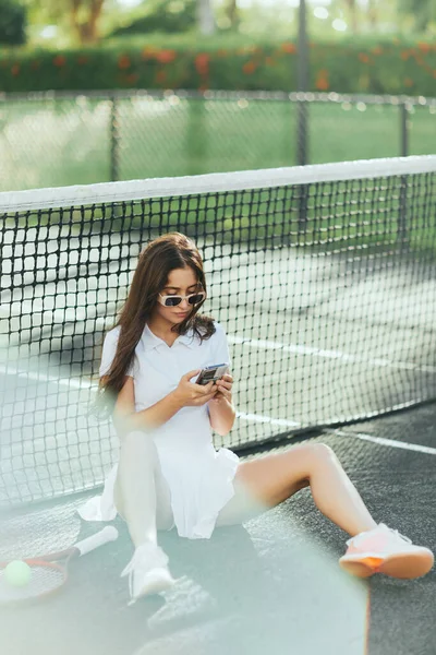 Court de tennis à Miami, jeune femme sportive aux cheveux longs assise en tenue blanche et lunettes de soleil tout en utilisant smartphone près de raquette et ballon, filet de tennis, fond flou, ville emblématique, été — Photo de stock