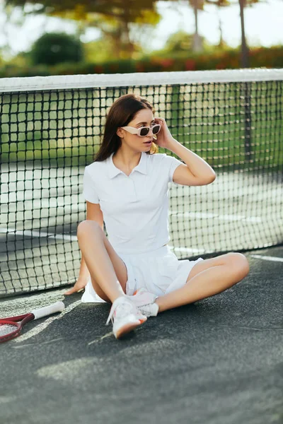 Relajación en la cancha de tenis en Miami, atlética joven con el pelo largo morena y mirando hacia otro lado mientras está sentado en traje blanco y gafas de sol cerca de raqueta, red de tenis, fondo borroso - foto de stock