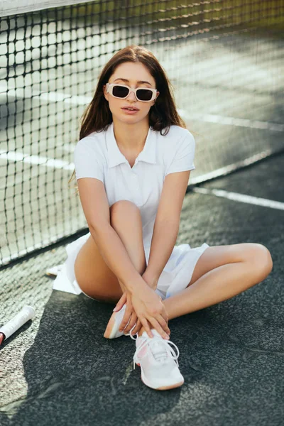 Tennis court in Miami, athletic young woman with brunette long hair sitting in white outfit, shoes and sunglasses near tennis net, blurred background, iconic city, physical activity — Stock Photo