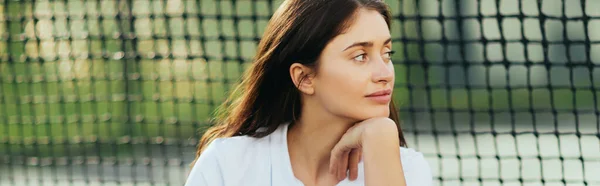 Joueuse assise sur un court de tennis, jeune femme intelligente aux cheveux longs bruns assise en tenue blanche près d'un filet de tennis, fond flou, Miami, regardant ailleurs, bannière — Photo de stock