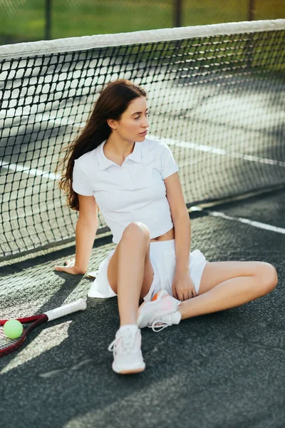 Jugadora de tenis descansando después del partido, mujer con pelo largo morena sentada en traje blanco cerca de raqueta con pelota y red de tenis en Miami, fondo borroso, ciudad icónica, pista de tenis, tiempo de inactividad - foto de stock