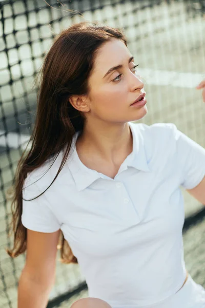 Portrait of attractive young woman with long brunette hair wearing white polo shirt and looking away after training on tennis court, tennis net on blurred background, Miami, Florida — Stock Photo