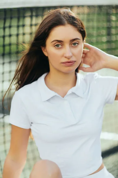 Porträt einer hübschen jungen Frau mit langen brünetten Haaren, die weißes Poloshirt trägt und nach dem Training auf dem Tennisplatz in die Kamera schaut, Tennisnetz auf verschwommenem Hintergrund, Miami, Florida — Stockfoto