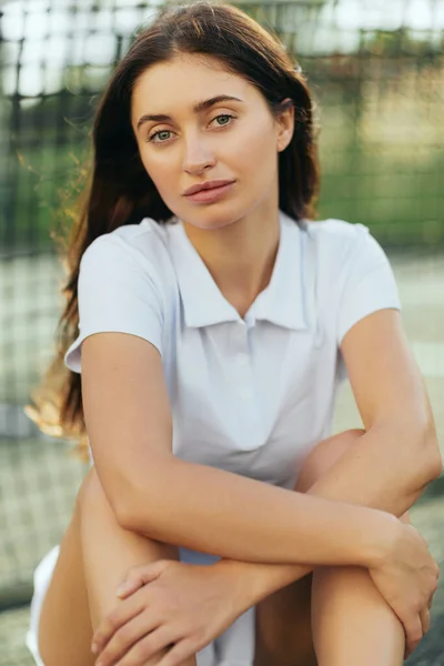 Cancha de tenis en Miami, retrato de una jugadora de tenis con el pelo largo morena vistiendo polo blanco y mirando a la cámara después del entrenamiento, red de tenis sobre fondo borroso, Florida - foto de stock