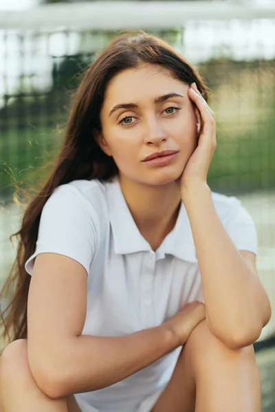 Tennis court, active lifestyle, portrait of female tennis player with brunette long hair wearing white polo shirt and looking at camera, tennis net on blurred background, Miami, Florida — Stock Photo